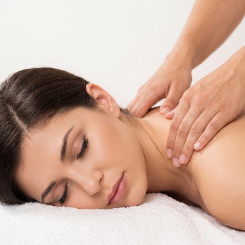 Kobieta z zamkniętymi oczami podczas masażu relaksacyjnego barków i szyi w salonie spa Warszawa. Zbliżenie na dłonie terapeuty wykonujące masaż antystresowy.