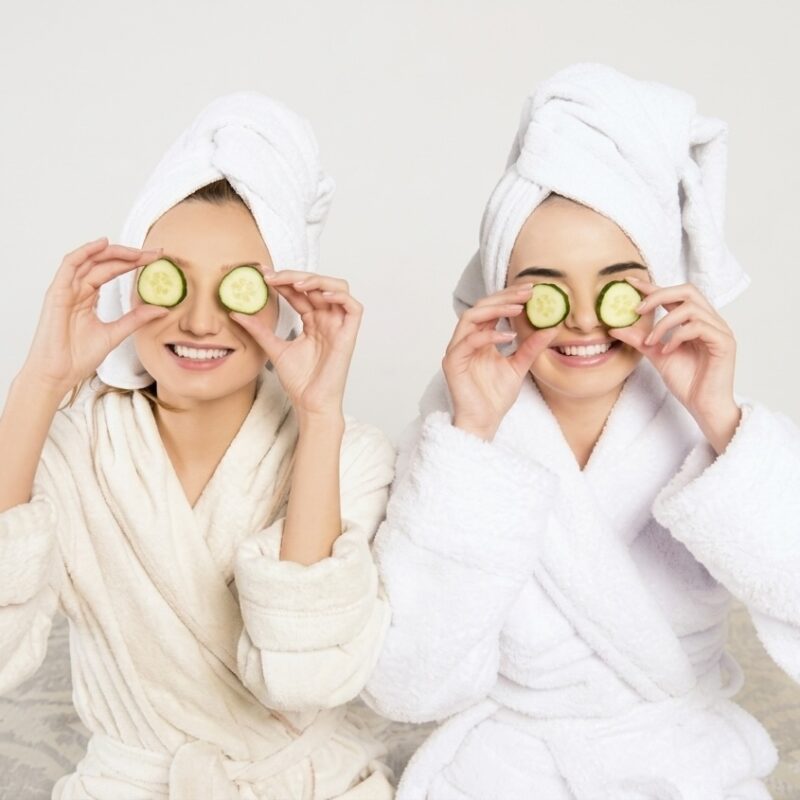 Dwie uśmiechnięte kobiety w białych szlafrokach z plastrami ogórka na oczach podczas warsztatów pro-aging w Alchemy Day Spa Warszawa.